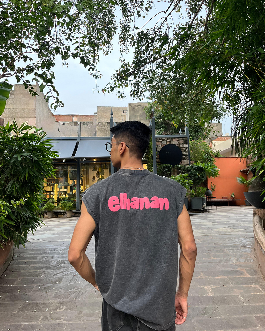 Person wearing a gray sleeveless shirt with 'elhanan' printed on the back, standing outdoors with greenery and buildings in the background.