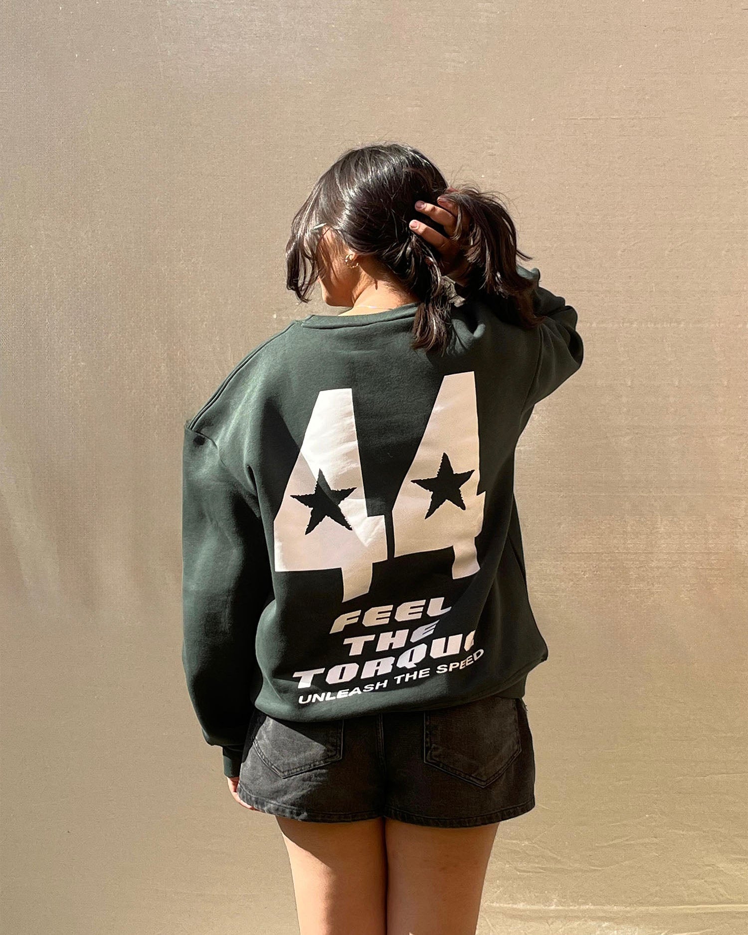 Person wearing a green sweatshirt with a graphic design on the back against a beige wall.